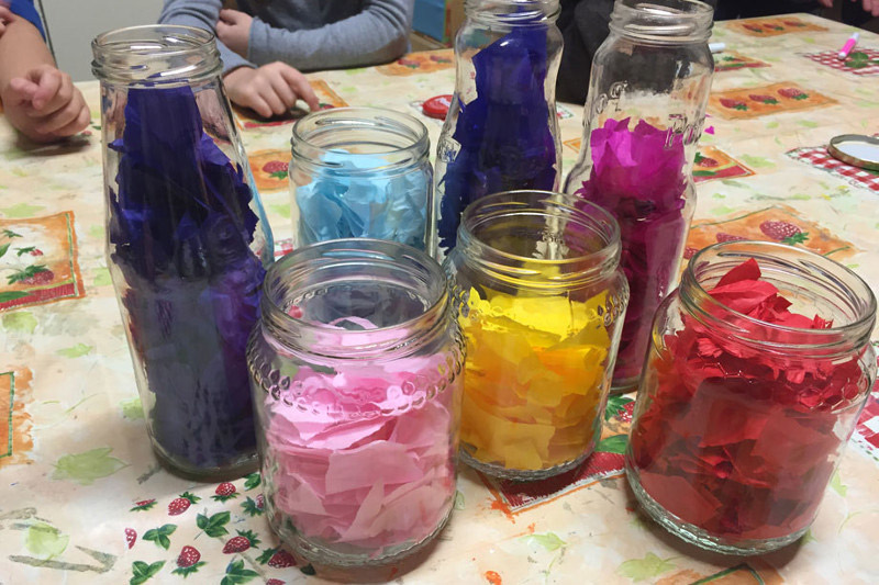 L’immagine mostra un insieme di
              vasetti di vetro contenenti dei pezzi di carta colorata. Ogni contenitore raccoglie pezzi di colore diverso